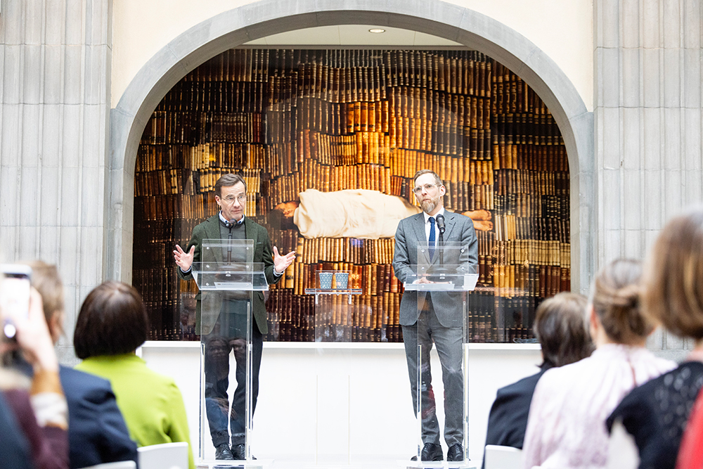 Två män står bakom talarstolar. En av dem håller tal. Bakom dem en tavla.