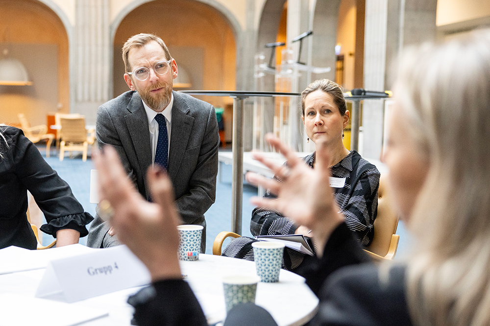 Personer sitter vid ett runt bord och pratar. Det står "Grupp 4" på en lapp på bordet.