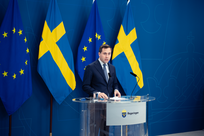 Benjamin Dousa står vid talarstol inför svenska flaggor och EU-flaggor.